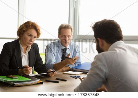 Business people taking interview while sitting at table in board room all together. Serious business workers listening to new worker sitting in front. Employee concept. Job concept.