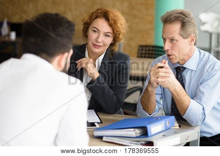 Business representatives communicating with young man and asking him to tell about his previous experience. Happy HR specialists of company taking interview.
