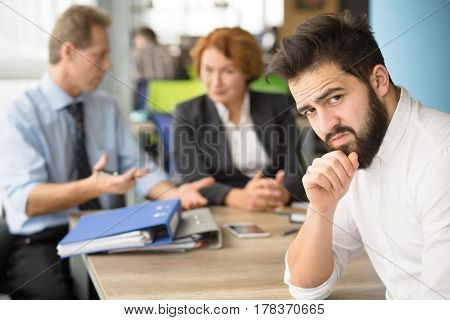 Interview concept. Young bearded applicant man having stress before interview while business employees communicating on background.