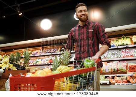 Image of young cheerful man with shopping trolley choosing fruits in supermarket. Looking aside.