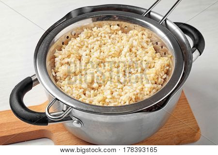 Colander with brown rice in saucepan on wooden board