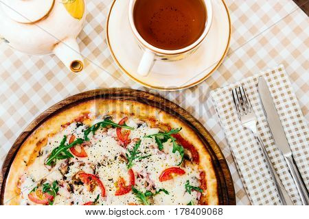 Pizza with cheese, tomato and meat, with cup of tee on served table with plaid tablecloth