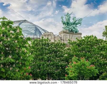 Bronze allegorical statue group over the southern wing of the main facade and part of the glass vaulted roof of the Great Palace built in 1900 with flowering chestnuts in the foreground in Paris