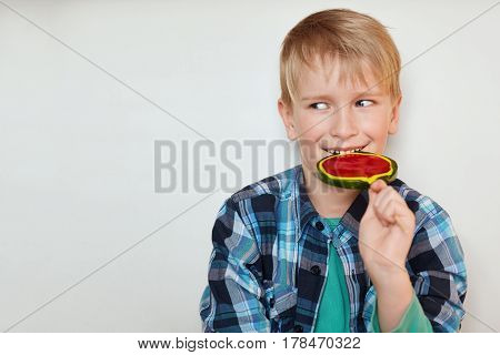 Cute blond boy with blue eyes dressed in checked shirt licking lollipop having happy expression looking aside on white background. Tiny kid looks aside enjoying his sweet candy with pleasure