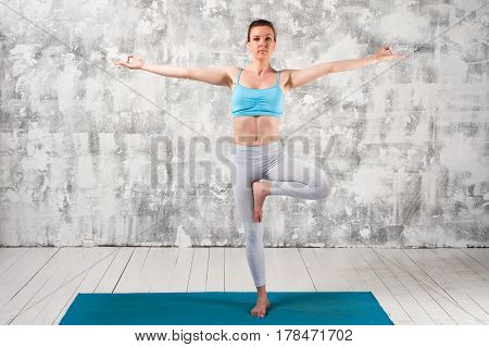 Young attractive woman wearing sportswear staying in Yoga Tree Pose. Fit girl exercising on blue mat against grey wall.
