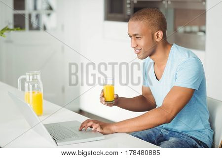 Smiling man using laptop and drinking orange juice in the ktichen