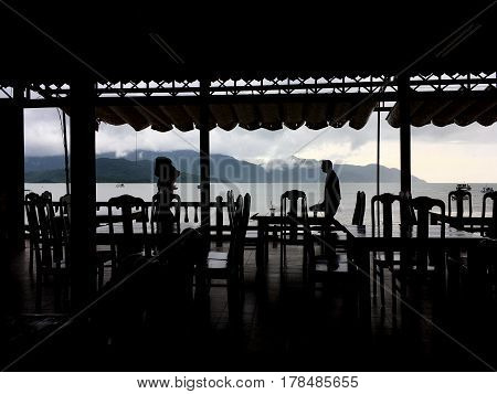 the view through the café to the sea, plenty of tables and chairs, subdued, the silhouettes, the roof in the form of a canopy above the café, the waiters at the background to see the sea, mountains with low clouds