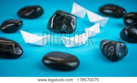 White paper boats in sigle file between abstract rock stones on blue background. Shot from Side Perspective.