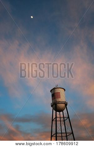 Half moon shines brightly in the pink and blue skies above a water tower with the American flag in Alamosa, Colorado at sunset