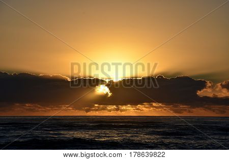 Beautiful sunrise cloudscape over ocean background. Sun rays beaming through picturesque clouds above sea. Blue sky with clouds sea and sun on the horizon. Beautiful sunrise over the quiet calm sea.
