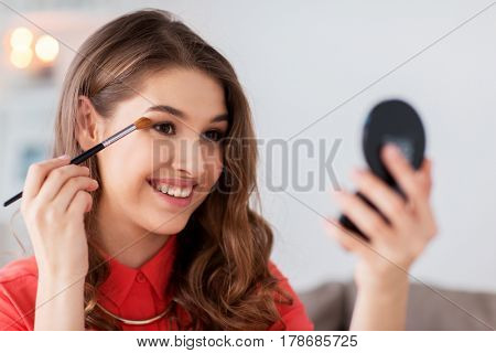 beauty, makeup, cosmetics and people concept - happy smiling young woman applying eyeshade with eye shadow brush and looking to mirror at home