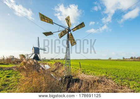 Small metal windmill and a large wooden hollow post mill together in a Dutch polder landscape on a sunny day in the fall season.