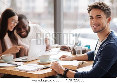 Studying together. Young handsome guy smiling while sitting at cafe on background of his studying fellow students.