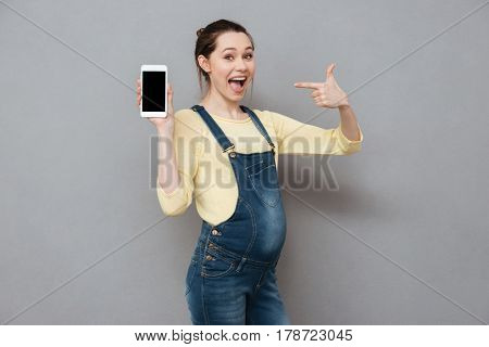 Portrait of a happy excited pregnant woman pointing finger at blank screen mobile phone isolated on a gray background