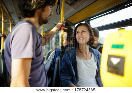 Young couple in inside of the city bus. The girl sits the fellow stands nearby. Young people look at each other and smile.
