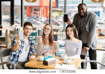 Our best helpmates. Happy international youngsters are showing off their modern smartphones while sitting at cafe.