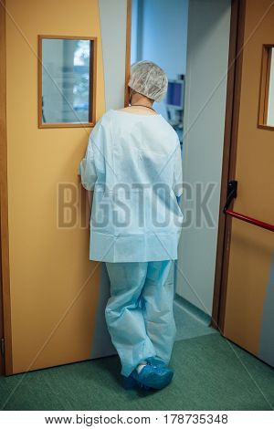 Surgeon dressed in one-off surgical uniform leans against the door and looks into the operating room