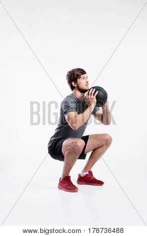 Handsome hipster fitness man in gray t-shirt holding medicene ball, doing squat. Studio shot on gray background.