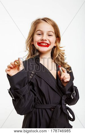 Cute little girl in coat copying mother, putting on red lipstick, smiling cheerfuly, looking straight.