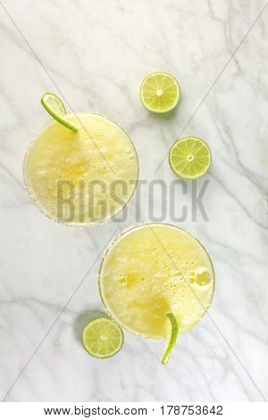 An overhead shot of two lemon Margarita cocktails with wedges of lime on a white marble table with copy space
