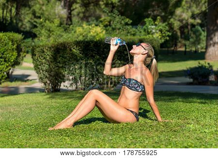 Attractive healthy young smiling beautiful curly blonde hair slim girl fashion portrait in black dress drinks water doing sport outdoor fitness diet & body care concept in background green grass meadow. Healthy woman lifestyle concept. Black sunglasses.