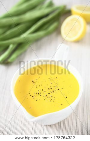 Hollandaise sauce a basic sauce of the French cuisine served in a sauce boat with ground black pepper on top lemon and raw green beans in the back photographed with natural light (Selective Focus Focus one third into the image)