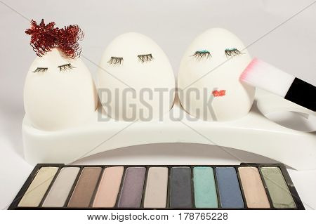 Three eggs with eyelashes in front of a palette of eye shadows make them make-up