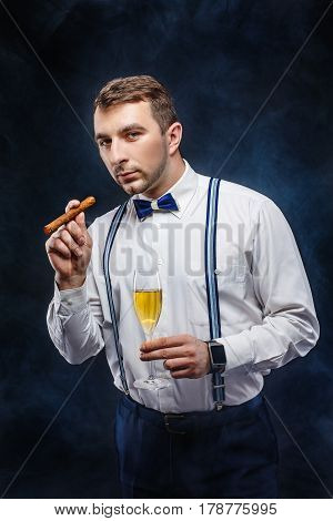 Portrait of a handsome man in bow-tie and suspenders holding glass of beverage and cigar while looking at camera. Smoke on background. Studio shot