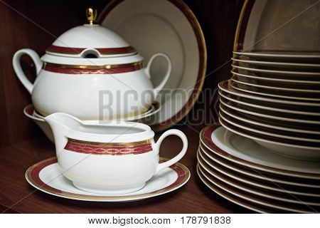 Closeup of white decorated china teaset and piled plates on wooden shelf