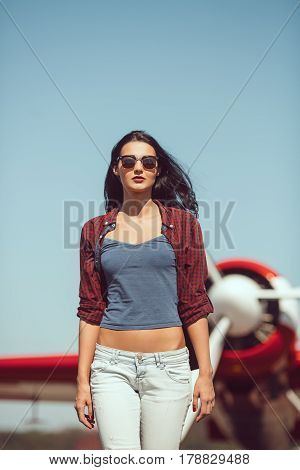 Pilot woman next to propeller plane outdoors in sunny day. Attractive young multi-racial Asian Caucasian sexy girl in jeans and shirt walking from sport airplane in airport.