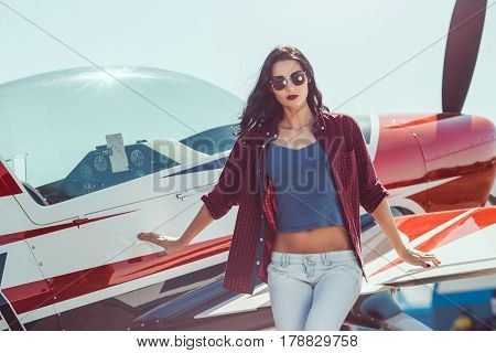 Pilot woman standing at cabin plane outdoors in sunny day. Attractive young multi-racial Asian Caucasian sexy girl in jeans and shirt near wing sport airplane in airport looking at camera.