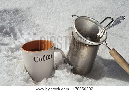 Cup of fresh made coffee next to a coffee maker and drainer. All placed on the snow.