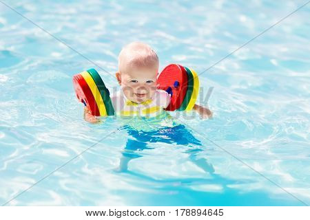 Happy laughing little baby boy playing in outdoor swimming pool on a hot summer day. Kids learn to swim. Child with colorful floaties. Swimming aid for kid. Family vacation in tropical resort.