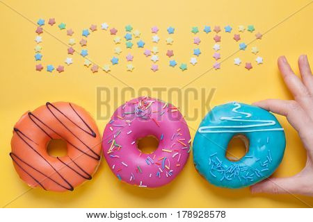 A female hand takes a blue donut in the glaze. Nearby lie pink and orange donuts. Top of the inscription donuts made of sugar stars.