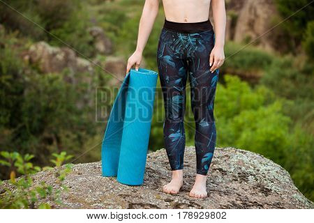 Young sportive girl holding yogamat, standing on rock in canyon. Copy space. Close up of legs.