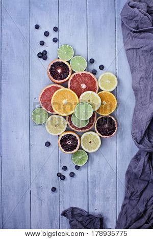 Variety of citrus fruits (orange blood oranges lemons grapefruits and limes) with blueberries with grey fabric over a blue rustic wood table top. Image shot from overhead.
