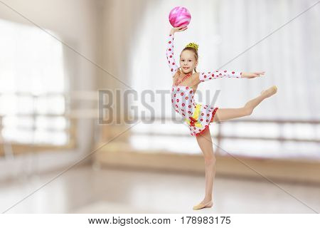 Beautiful little girl gymnast dressed in sports suits for competition, performs exercises with the ball.In the sports hall with mirror and a large semi-circular window.