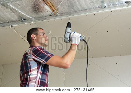 Young Man In Goggles Fixing Drywall Suspended Ceiling To Metal Frame Using Electrical Screwdriver On