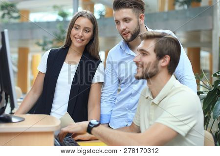 Group Of Young People In Casual Wear Sitting At The Office Desk And Discussing Something While Looki