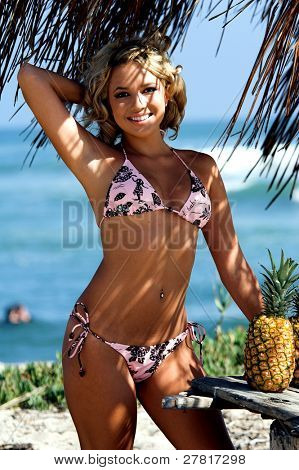 Ragazza bionda sexy del bikini in spiaggia in bikini rosa e marrone di Swimbay sotto un ombrello palapus w