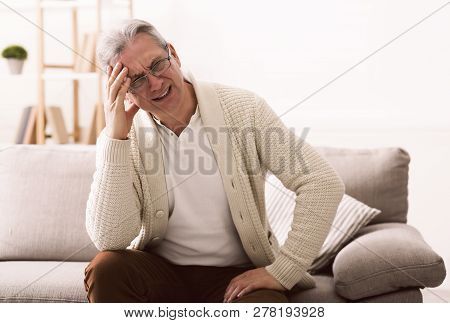 Senior Man Suffering From Headache, Sitting On Sofa At Home