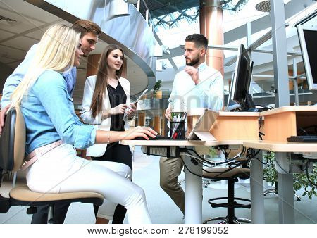 Group Of Young People In Casual Wear Sitting At The Office Desk And Discussing Something While Looki