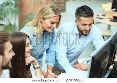 Group Of Young People In Casual Wear Sitting At The Office Desk And Discussing Something While Looki