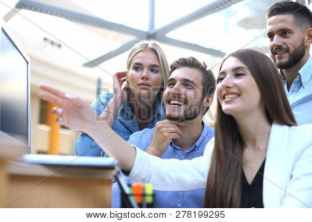Group Of Young People In Casual Wear Sitting At The Office Desk And Discussing Something While Looki