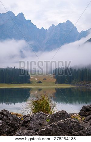 View Over Lake Laghi Di Fusine On A Foggy Morning With Some Rocks At The Beach And Mountain Range Ma