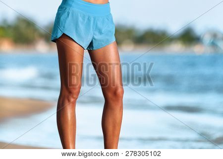 Woman legs fit girl wearing blue shorts standing walking on beach. Lower body crop of toned and tanned legs. Attractive healthy active runner person.