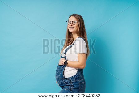 Pregnant Woman Caressing Her Belly Over Blue Background. A Cute Young Woman Wearing Glasses, And Den