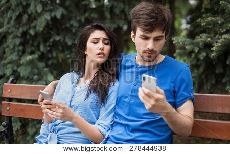 Young Couple In The Park. Boy Is Checking His Phone Hiding It. Girl Is Lookin Into Boyfriends Phone 