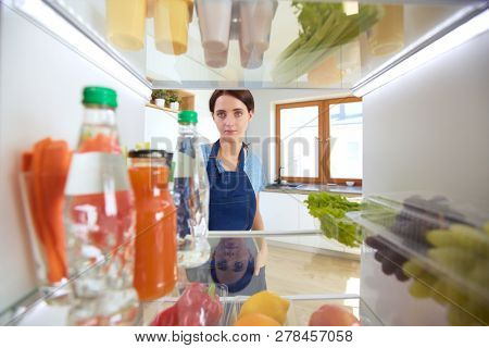 Portrait of female standing near open fridge full of healthy food, vegetables and fruits. Portrait of female