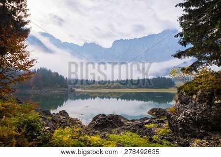 View Over Lake Laghi Di Fusine On A Foggy Morning With Some Rocks At The Beach And Mountain Range Ma